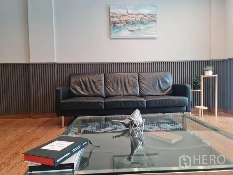 Townhouse for sale in Suan Luang, Bangkok - Modern living room with black leather sofa, glass coffee table, wood floor and wall art.