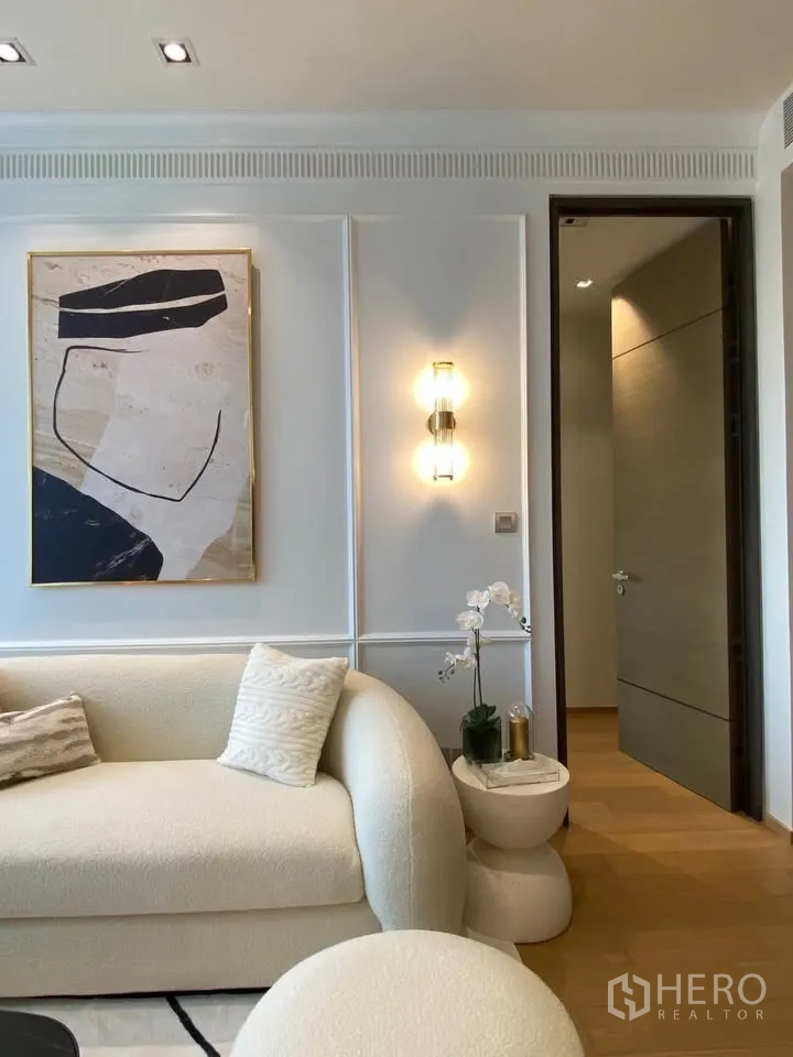 Condominium for rent in Pathum Wan, Bangkok - Living room corner with cream sofa, wall art, sconce lighting, and a small side table with orchid.