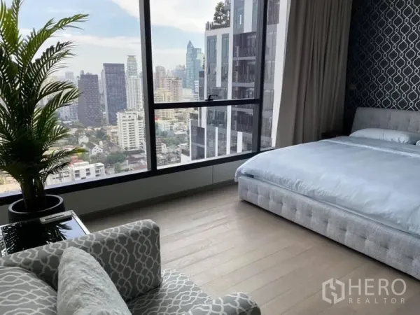 Condominium for rent or sale in Watthana, Bangkok - Master bedroom with floor‑to‑ceiling windows, upholstered bed and armchair overlooking the Bangkok skyline.