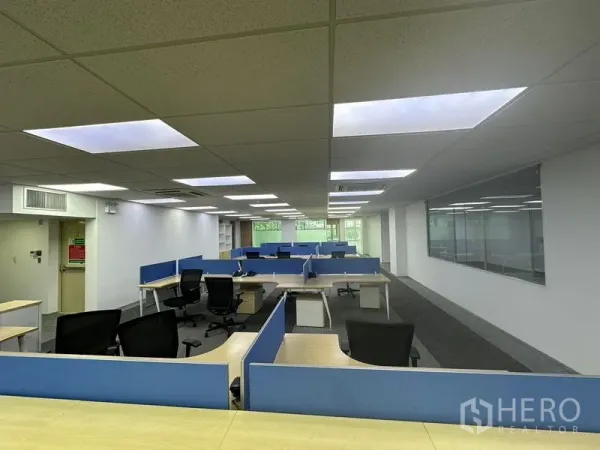 สำนักงาน ให้เช่า - โต๊ะทำงานแบบโมดูลาร์เรียงหลายชุดใต้ไฟฝ้า พร้อมผนังกระจกและวิวต้นไม้ด้านหน้า
