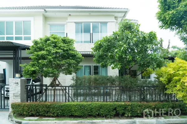 บ้าน ให้เช่าหรือขายในSaphan Sung, Bangkok - ด้านหน้าบ้านพร้อมหน้าต่างบานเลื่อน ต้นไม้ร่มรื่น พุ่มไม้ตัดแต่ง และรั้วเหล็กสีดำ