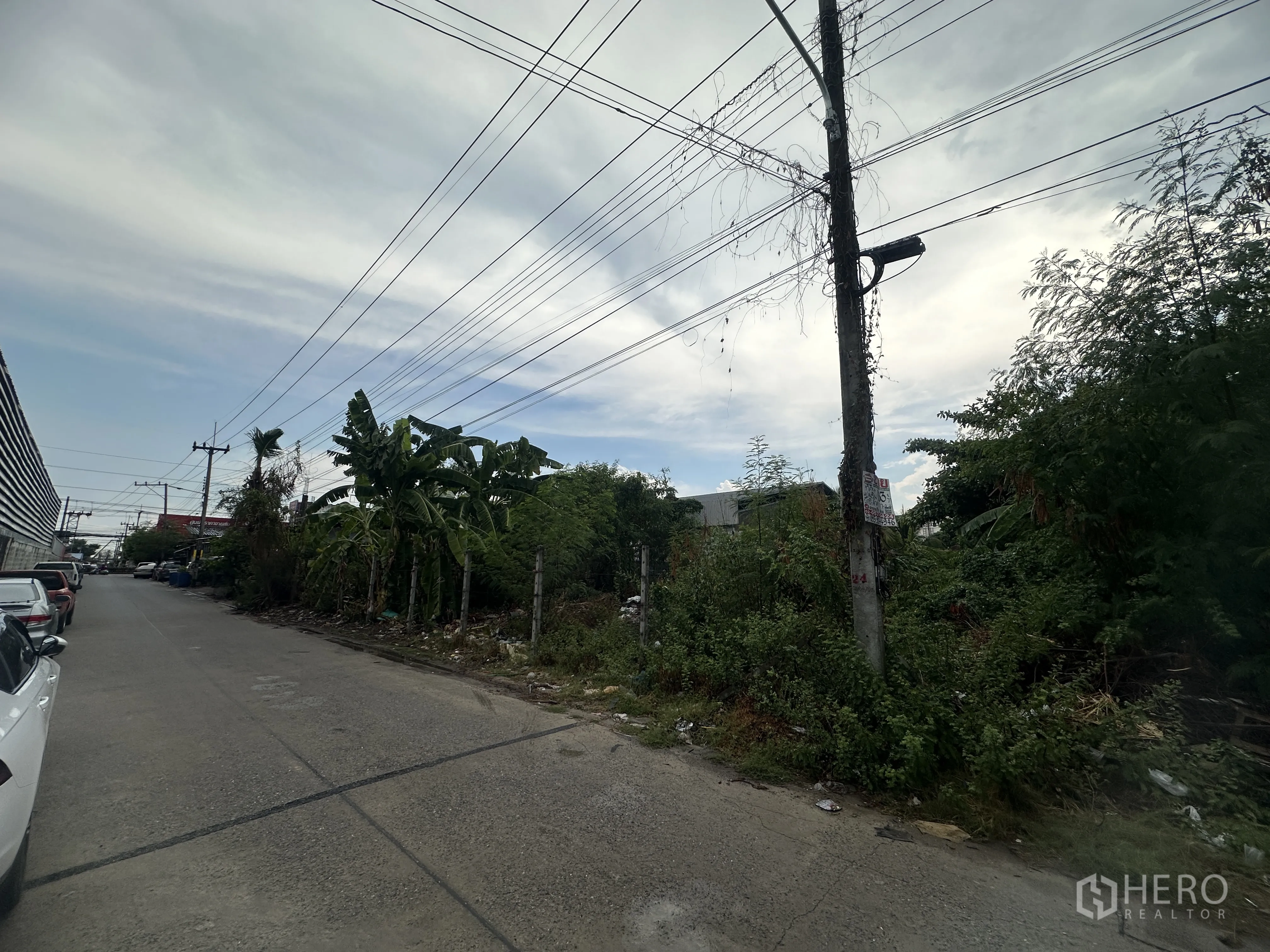 土地 for sale in Lam Luk Ka, Pathum Thani - 社区混凝土道路与架空电线旁的一块杂草丛生的空地。