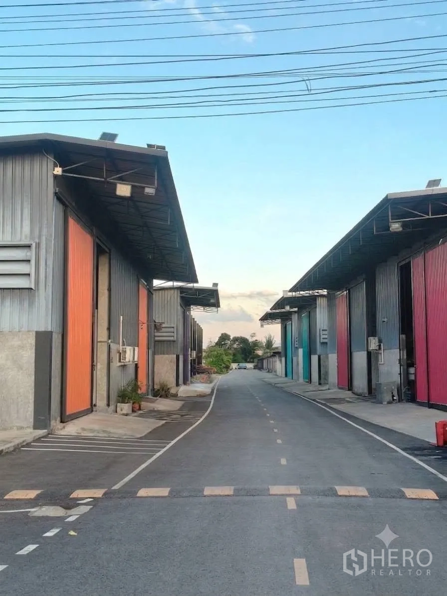 Warehouse for rent in Bang Bua Thong, Nonthaburi - Paved internal estate road lined with warehouse units, overhead power lines, and a speed bump.