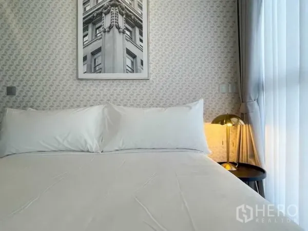Condominium for rent in Ratchathewi, Bangkok - Close-up of a cozy bed against patterned wallpaper with a brass bedside lamp and sheer curtains.