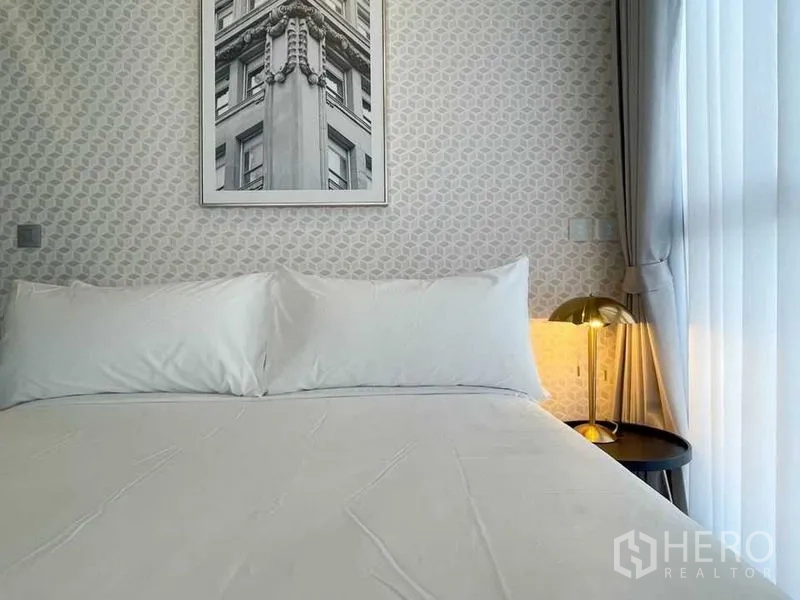 Condominium for rent in Ratchathewi, Bangkok - Close-up of a cozy bed against patterned wallpaper with a brass bedside lamp and sheer curtains.