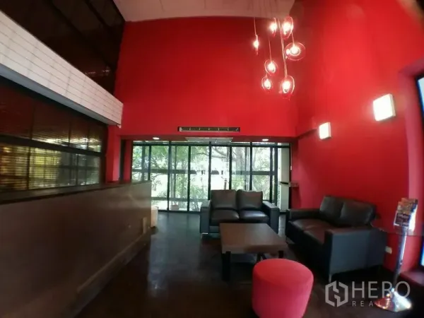 办公空间 出租在Phra Khanong, Bangkok - 双挑高红色墙面休闲区，皮沙发与吊灯，外面是绿景。