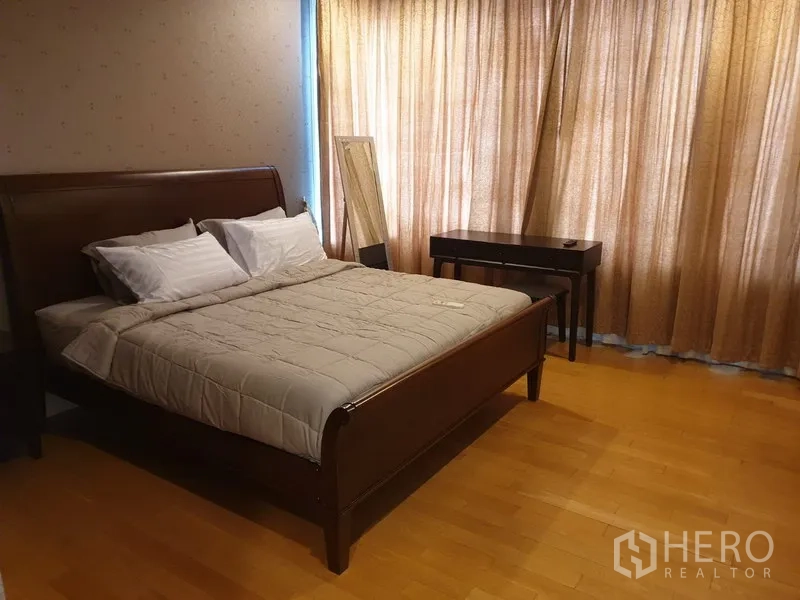 Condominium for rent in Khlong Toei, Bangkok - Spacious master bedroom with wooden bedframe, desk, wood floors, and full-length curtains.