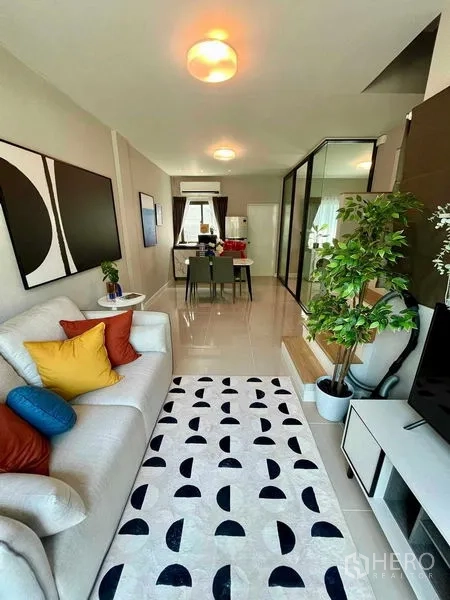 Townhouse for sale in Sai Mai, Bangkok - Cozy living room with sofa, TV console, patterned rug, and outlook to the garden.