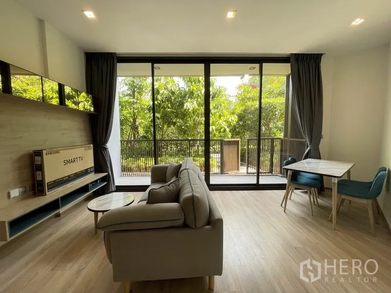 คอนโด ขายในWatthana, Bangkok - ห้องนั่งเล่นสว่าง โซฟาสีเทา ชุดโต๊ะทานอาหาร และหน้าต่างสูงจรดเพดานเปิดสู่ระเบียงวิวต้นไม้