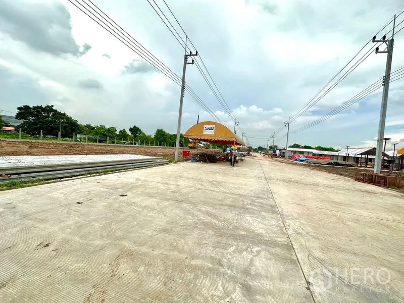 Land for sale in Lam Luk Ka, Pathum Thani - Concrete access road through an industrial development with utility poles, tents and stacked construction materials.