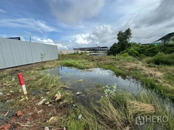 Land for sale in Samut Prakan - Open plot with a waterlogged area, metal fencing and greenery, with industrial buildings in the distance in Samut Prakan.