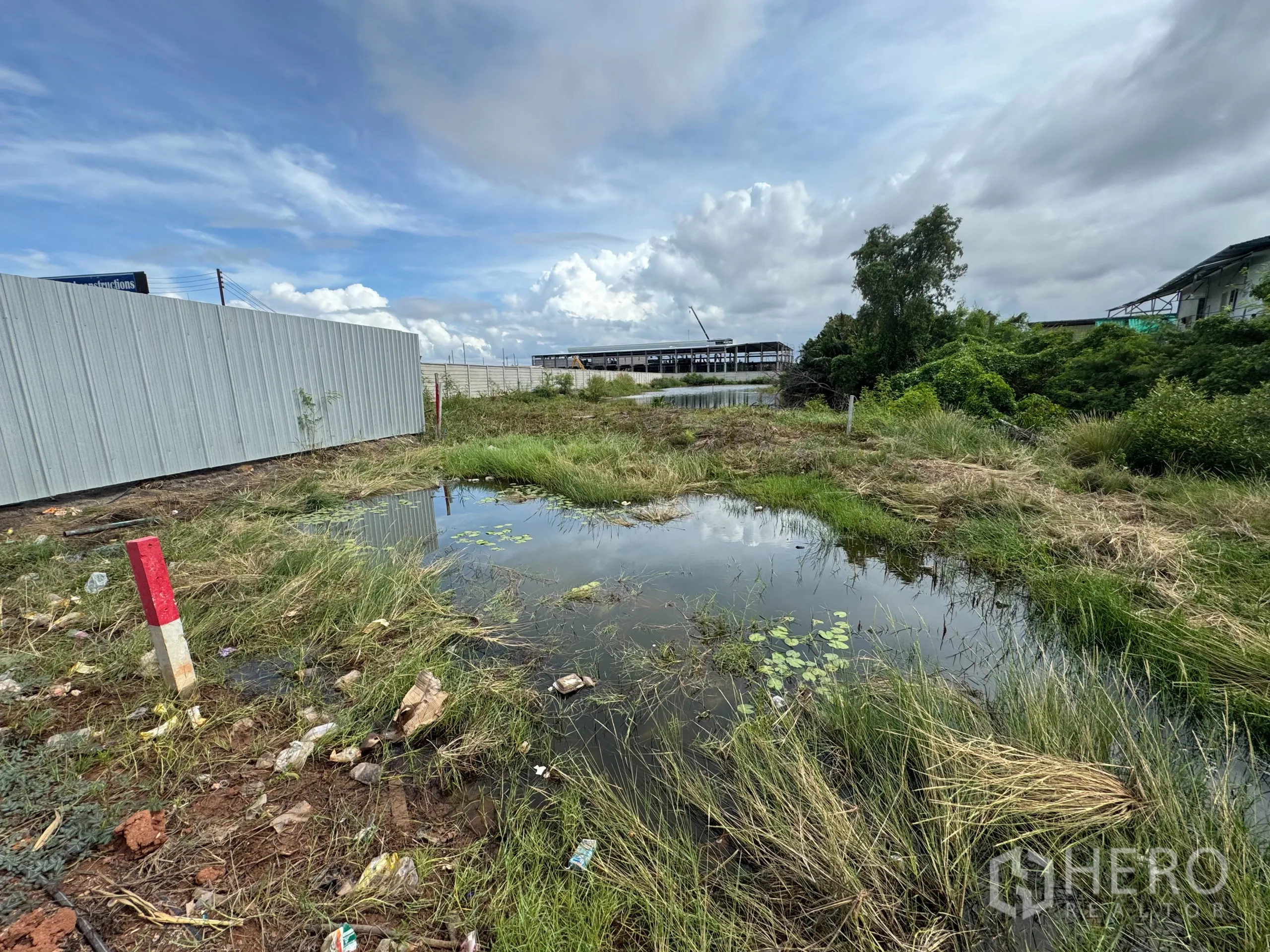 Land for sale in Samut Prakan - Open plot with a waterlogged area, metal fencing and greenery, with industrial buildings in the distance in Samut Prakan.
