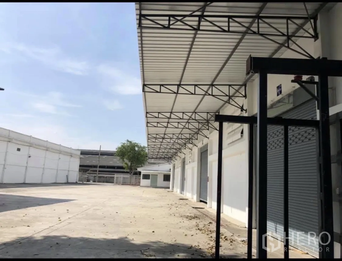 Warehouse for rent in Mueang Pathum Thani, Pathum Thani - Long covered walkway with multiple roller shutters and a broad maneuvering area in front.