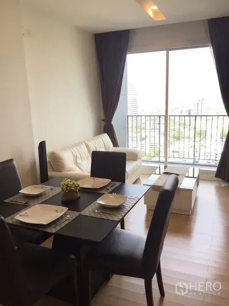 公寓 出租在Khlong Toei, Bangkok - 餐区连接明亮客厅，沙发与落地窗，通向可观城市景的阳台。