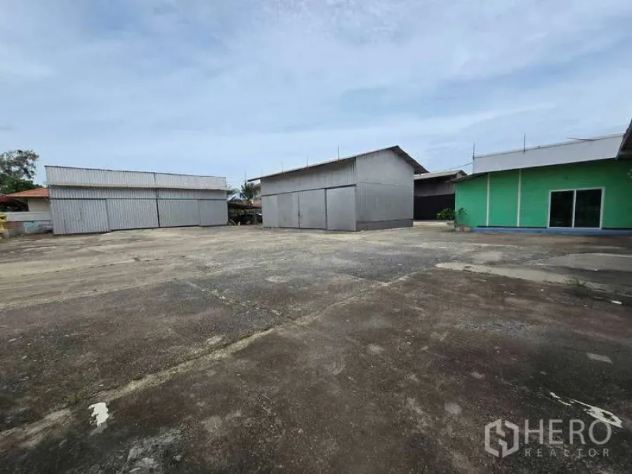 Warehouse for rent in Khlong Sam Wa, Bangkok - Spacious concrete yard with multiple metal warehouse sheds and a green office building.