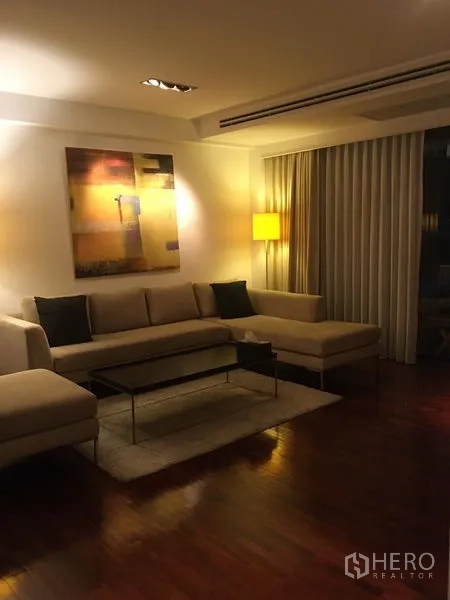 คอนโด ให้เช่า - ห้องนั่งเล่นสบายๆ โซฟาเข้ามุม โคมไฟตั้งพื้น งานศิลปะ และผ้าม่านสูงจรดเพดาน