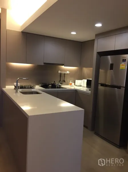 Condominium for rent in Watthana, Bangkok - Contemporary U-shaped kitchen with white countertop island, built-in cabinets and stainless refrigerator.