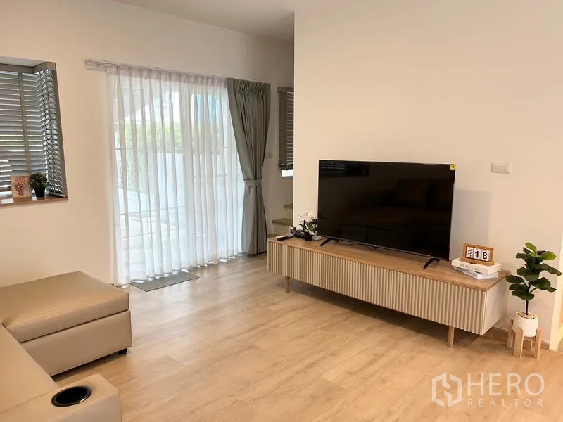 ทาวน์เฮาส์ ให้เช่า - ห้องนั่งเล่นมีชั้นวางทีวี ประตูกระจกเปิดสู่เฉลียง และผ้าม่านยาว.