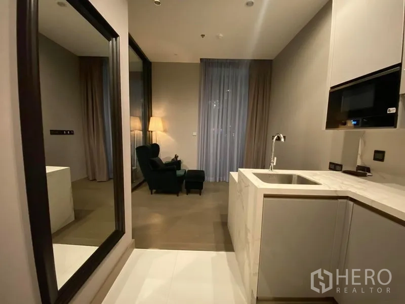 Condominium for rent in Huai Khwang, Bangkok - Open-plan living with marble L-shaped kitchenette, large wall mirror, and lounge chair by a floor-to-ceiling window.