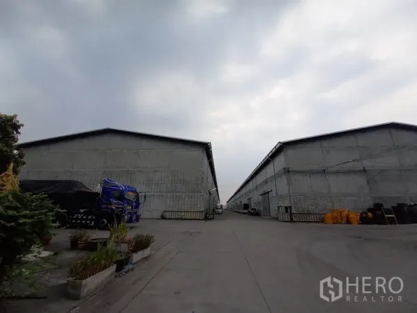 Warehouse for rent in Mueang Pathum Thani, Pathum Thani - Wide driveway between large concrete warehouse blocks in a private compound, showing ample truck access under a cloudy sky.