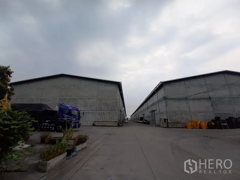 Warehouse for rent in Mueang Pathum Thani, Pathum Thani - Wide driveway between large concrete warehouse blocks in a private compound, showing ample truck access under a cloudy sky.
