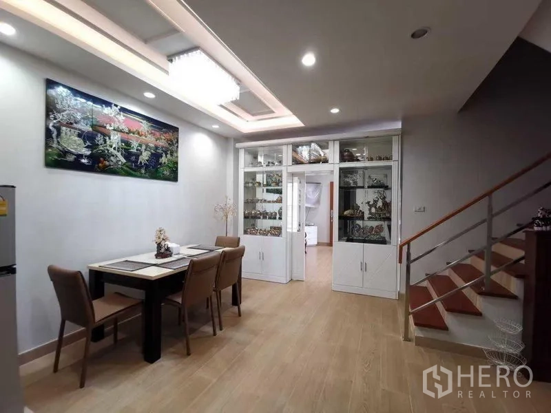 Detached House for rent in Prawet, Bangkok - Dining area with four‑seat table, recessed ceiling lights, built‑in glass cabinets and staircase.