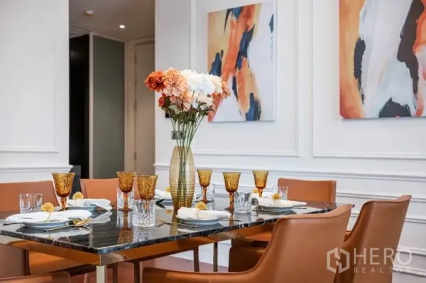 คอนโด ให้เช่าหรือขายในWatthana, Bangkok - โต๊ะอาหารหินอ่อนจัดเต็มสำหรับหกที่นั่ง มีแก้วสีอำพันและแจกันดอกไม้ หน้ากำแพงบัวและงานศิลป์