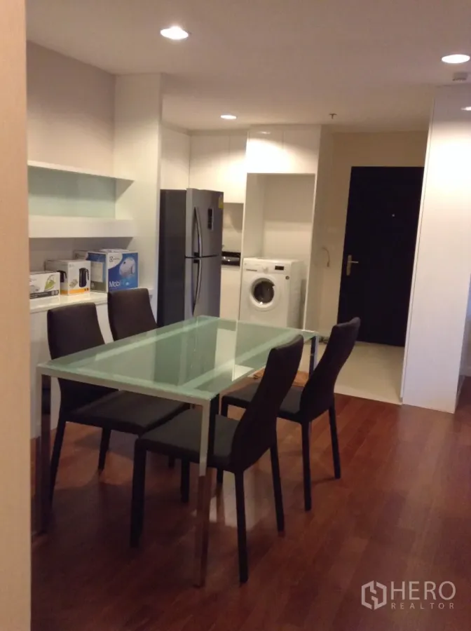 Condominium for rent in Huai Khwang, Bangkok - Open-plan dining and kitchenette with a glass table, four chairs, refrigerator and washing machine on warm wood flooring.