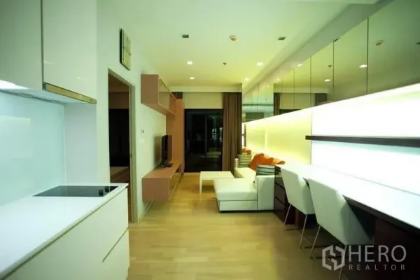 Condominium for rent or sale in Watthana, Bangkok - Open-plan living and kitchen featuring built-in desk, mirrored wall and sliding door to the balcony.