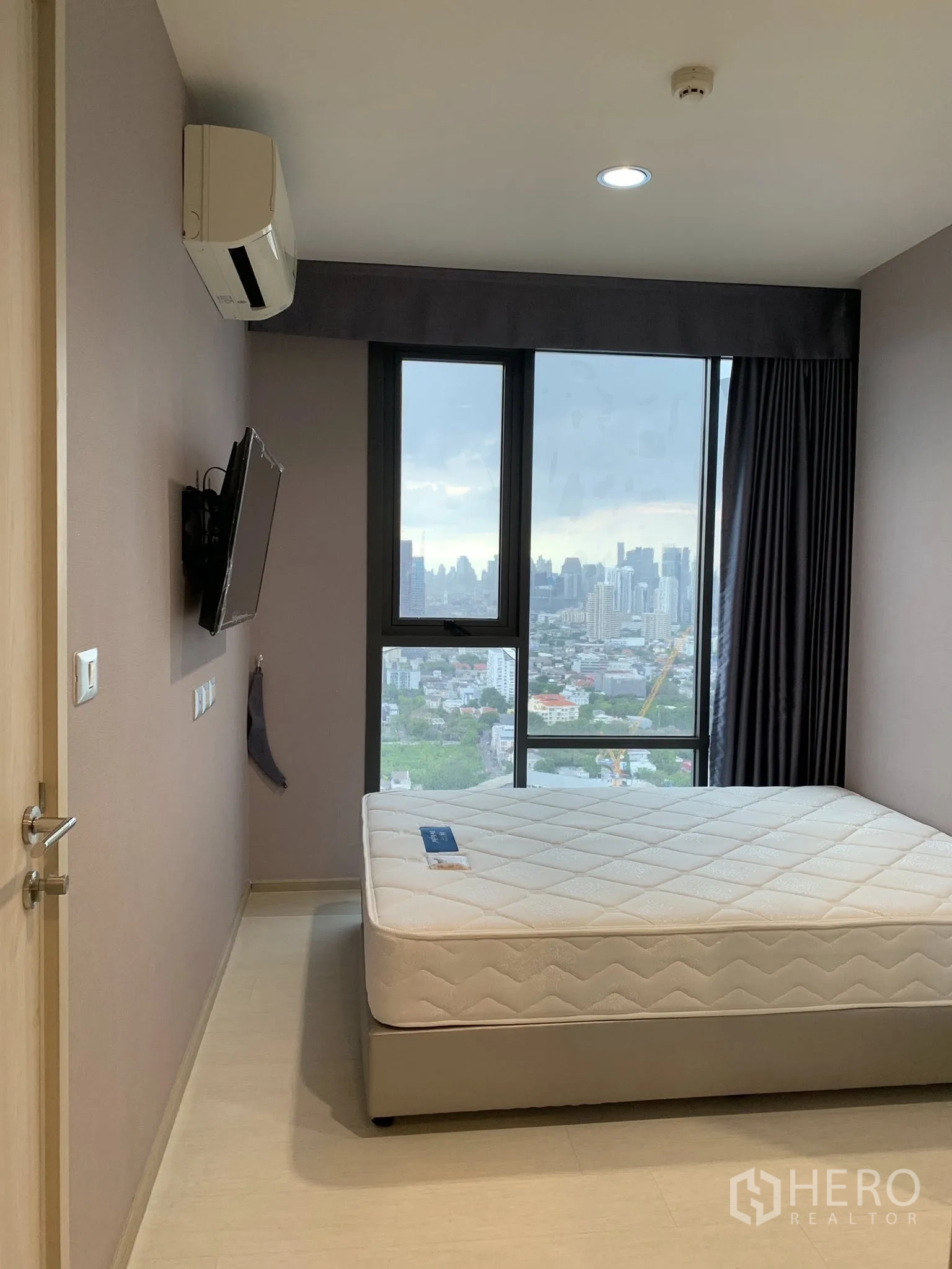 Condominium for rent in Khlong Toei, Bangkok - Bedroom with bed, wall-mounted TV, air conditioner, and window overlooking the city.