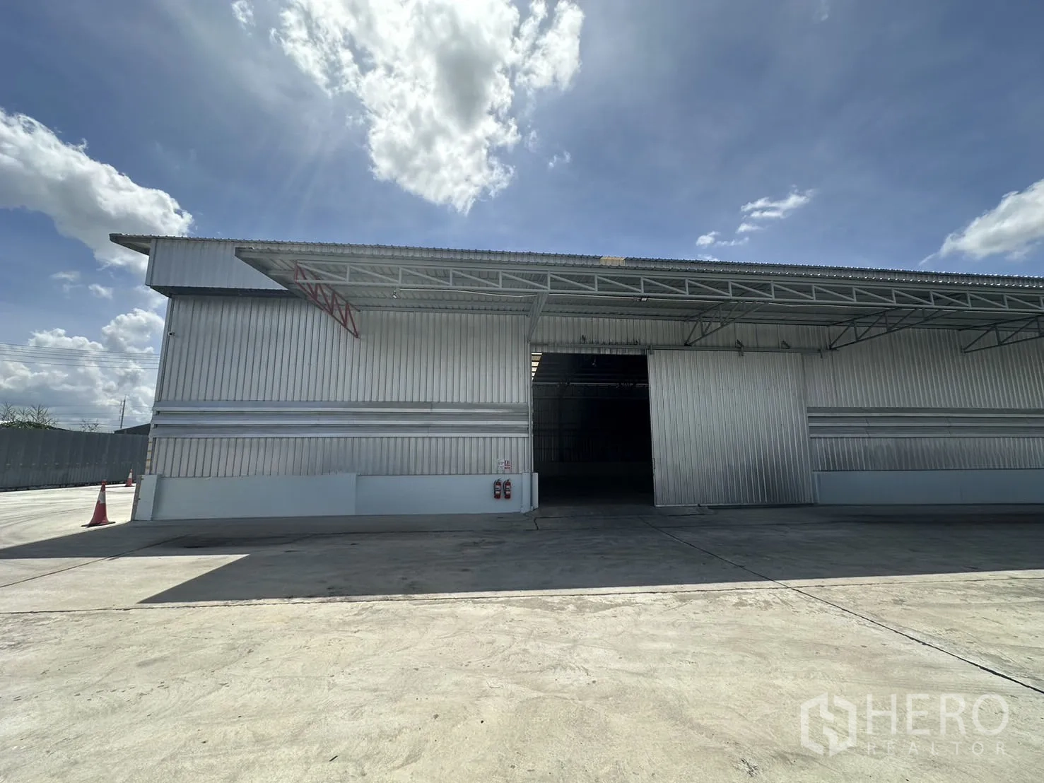 Warehouse for rent in Mueang Pathum Thani, Pathum Thani - Front exterior of a metal-clad warehouse with large sliding door, covered loading bay and wide concrete yard under a blue sky.