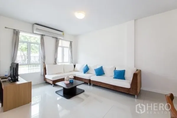 Townhouse for rent in Watthana, Bangkok - Bright living room with L-shaped sofa, air conditioning and large windows overlooking the front courtyard.