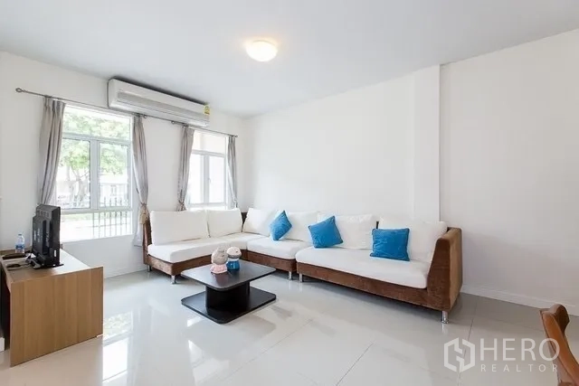Townhouse for rent in Watthana, Bangkok - Bright living room with L-shaped sofa, air conditioning and large windows overlooking the front courtyard.