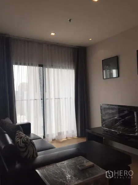 Condominium for rent in Khlong Toei, Bangkok - Bright living room with floor-to-ceiling windows, sheer curtains, balcony access, leather sofa and TV console.