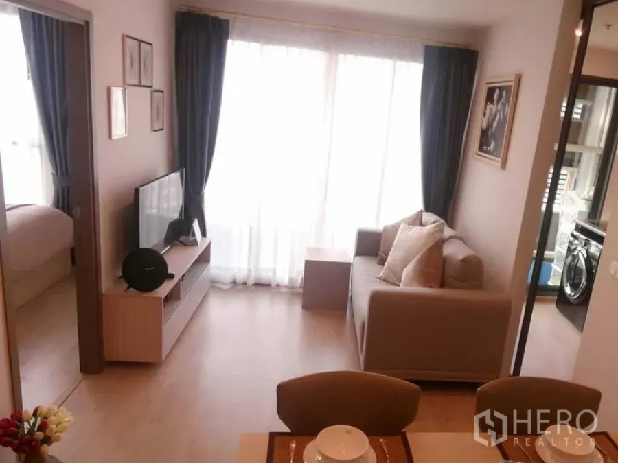 คอนโด ให้เช่าในBang Na, Bangkok - มุมนั่งเล่นรับแสงธรรมชาติ โซฟา ชั้นวางทีวี ผ้าม่านโปร่ง มองเห็นครัวและห้องนอนในคอนโดย่านบางนา.