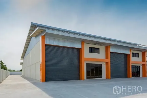 Factory for sale in Bang Sao Thong, Samut Prakan - Modern factory exterior with orange columns, metal roof, large roller shutters and a wide concrete yard.