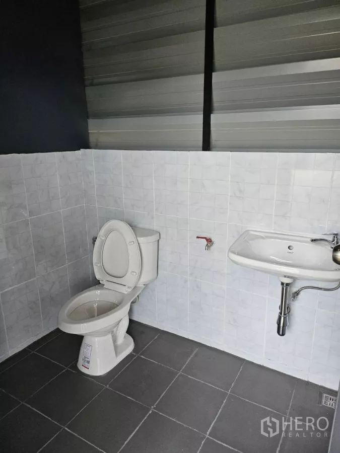 Warehouse for rent in Wang Thonglang, Bangkok - Second warehouse restroom with ceramic toilet, washbasin, and gray floor tiles.