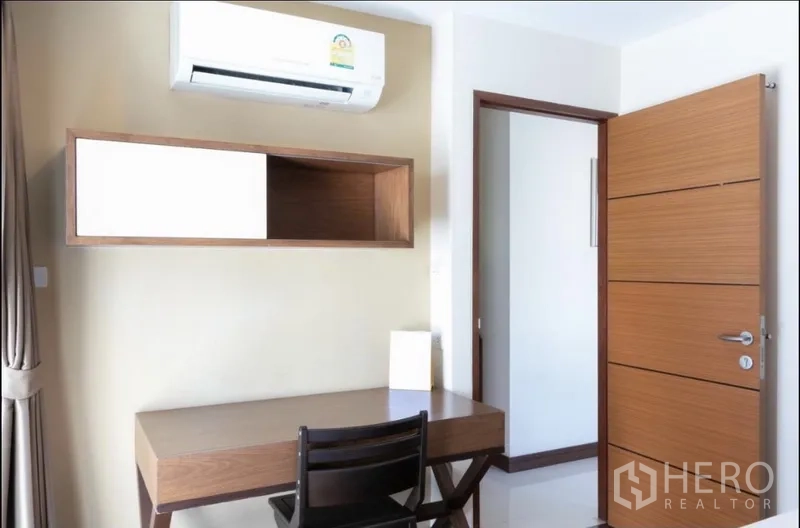 房间 出租在Bang Rak, Bangkok - 紧凑工作角，书桌、墙柜与空调，旁有木门。