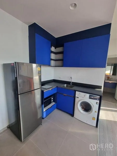 公寓 出租在Watthana, Bangkok - 紧凑厨房，蓝色柜体、黑色台面，配不锈钢冰箱、烤箱与洗衣机。