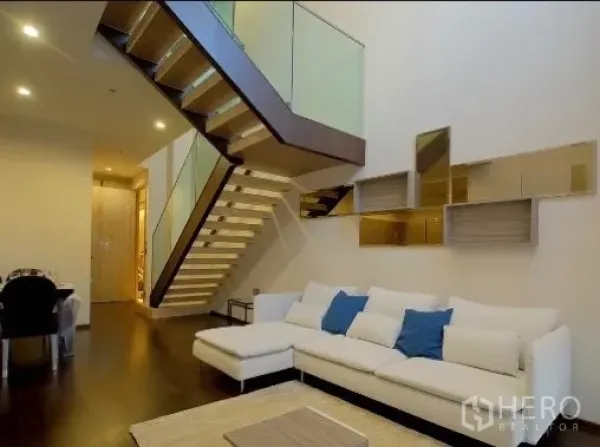 公寓 出租在Watthana, Bangkok - 挑高客厅的双层公寓，白色转角沙发与玻璃扶手楼梯。