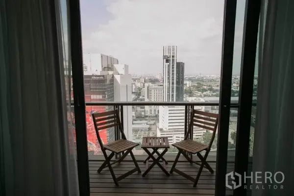 Condominium for rent or sale in Watthana, Bangkok - High-floor balcony with two wooden chairs and a table overlooking Bangkok skyline.