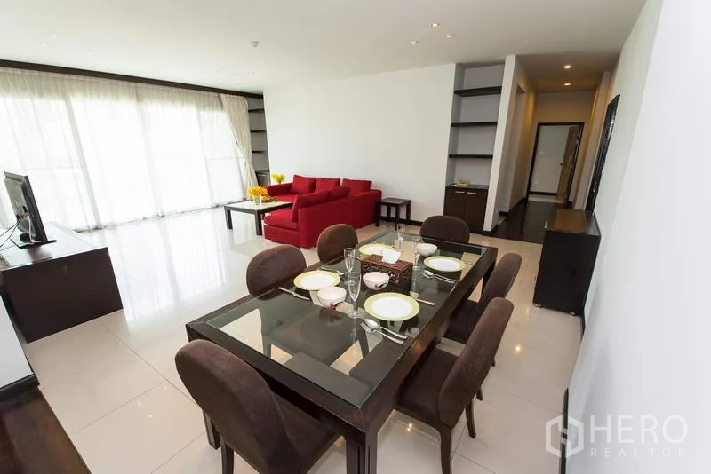 Apartment for rent in Sathon, Bangkok - Dining area with glass table for six facing a bright lounge and full-height curtains.