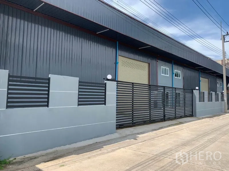 Factory for rent in Khlong Luang, Pathum Thani - Modern factory facade with grey metal cladding, rolling door and gated fence on a wide street.