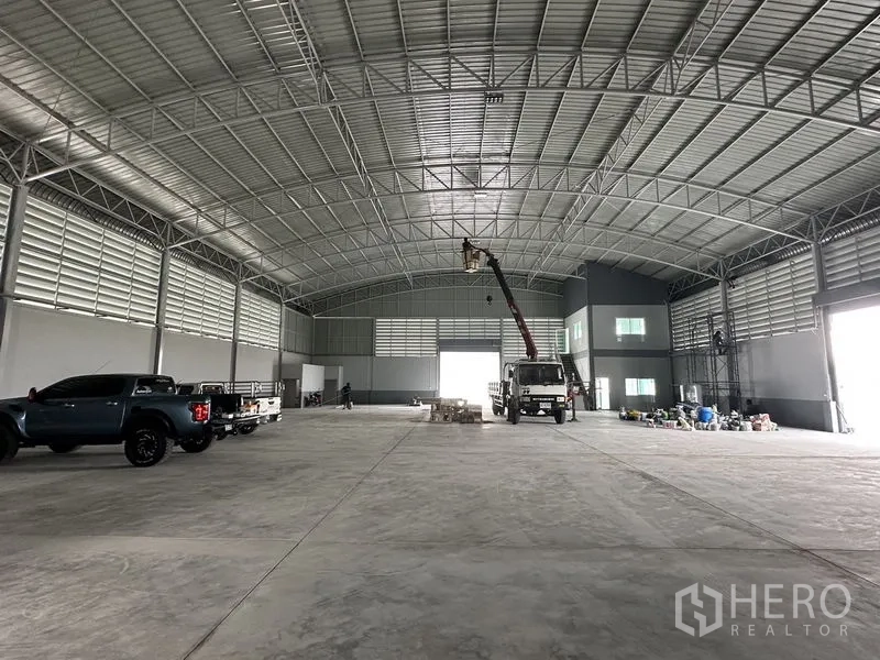 Factory for rent in Pathum Thani - Spacious factory floor under arched steel roof with trucks parked and bright end opening.
