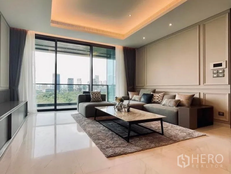 Condominium for rent in Pathum Wan, Bangkok - Spacious living room with L-shaped sofa, cove lighting, and floor-to-ceiling glass doors opening to skyline views.