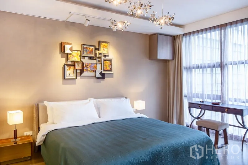 公寓 出租在Watthana, Bangkok - 舒适卧室，特大床、装饰画与枝形吊灯，落地窗旁设有书桌。