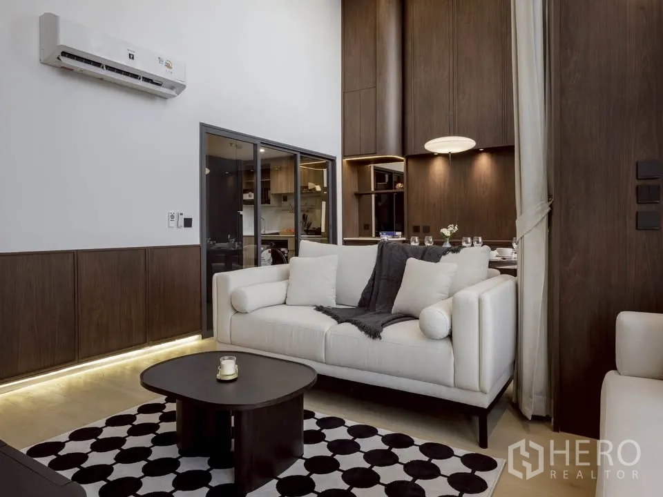 Condominium for rent in Khlong Toei, Bangkok - Cozy living area with white sofas, patterned rug, and glass-partitioned kitchen.