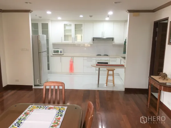 Condominium for rent in Watthana, Bangkok - Open-plan white kitchen with breakfast bar, glass-front cabinets and dining area on wooden flooring.