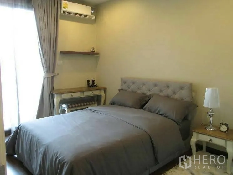 Condominium for rent or sale in Huai Khwang, Bangkok - Second bedroom with tufted headboard, bedside tables, writing desk and curtains to a balcony.