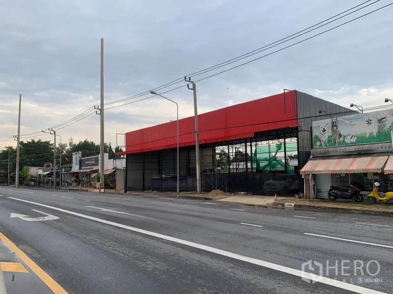 คลังสินค้า ให้เช่าในBueng Kum, Bangkok - มุมมองเฉียงของโกดังแผงสีแดงริมถนนหลายช่องจราจร พร้อมเสาไฟและทางเท้า.